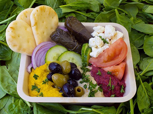 A greek salad is arranged in a compostable dish on top of a bed of spinach
