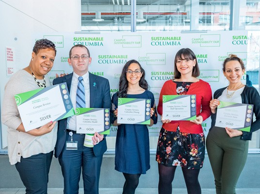 Green Leaders from Campus Services hold their certificates from the Workspace Certification program in front of a backdrop with the Sustainable Columbia logo.